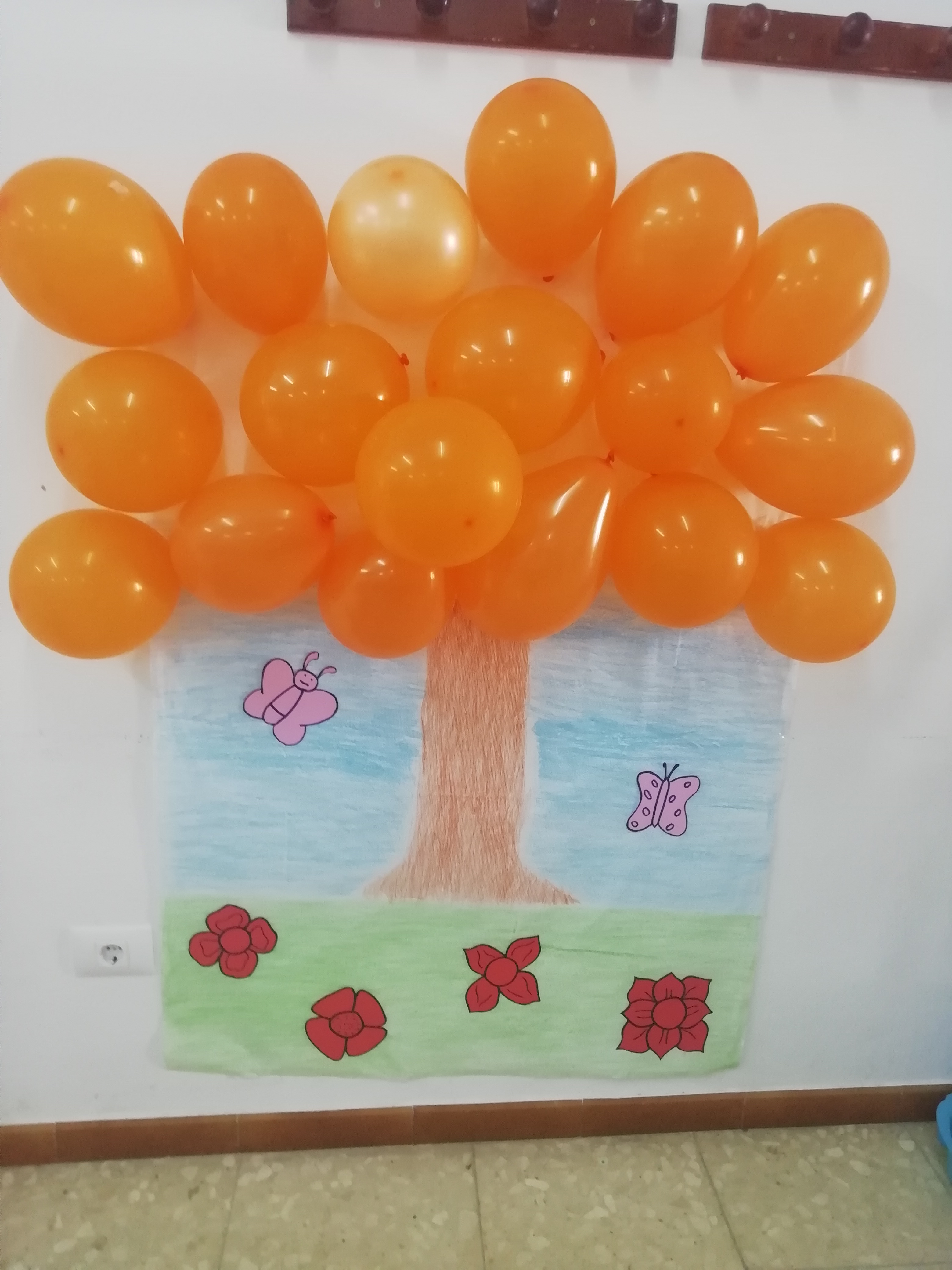 Mural de arbol pintado por niños y globos naranjas pegados en sus ramas.